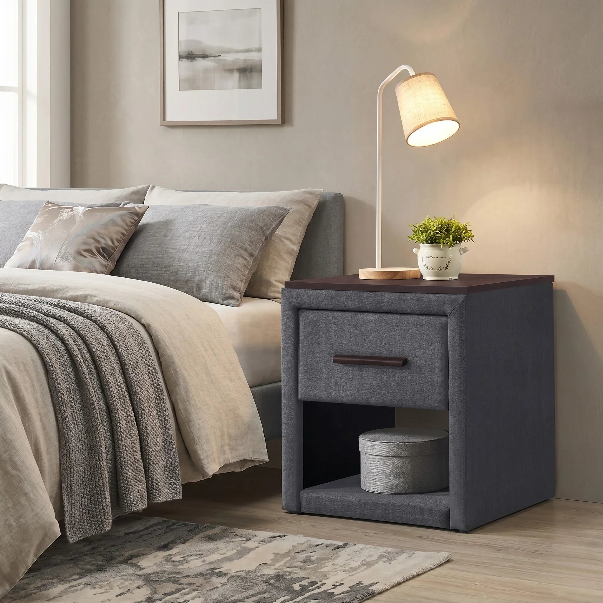 Gray upholstered nightstand next to a bed with pillows and a lamp in a bedroom setting.