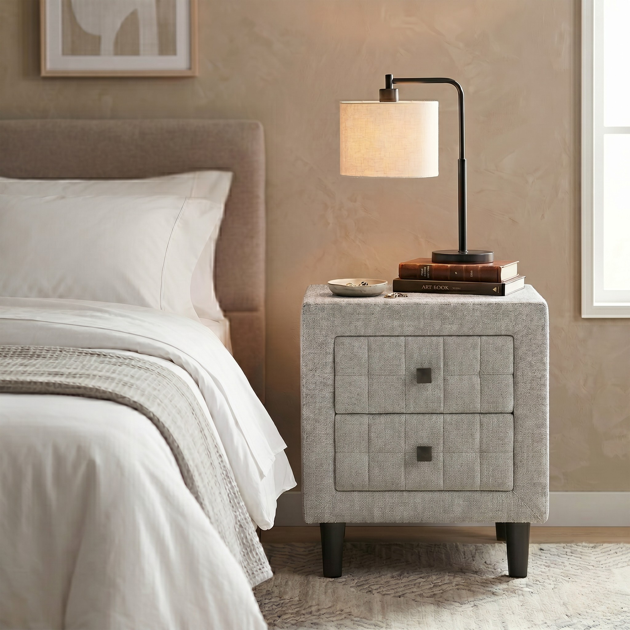 Gray upholstered nightstand with lamp and books next to a bed in a bedroom.