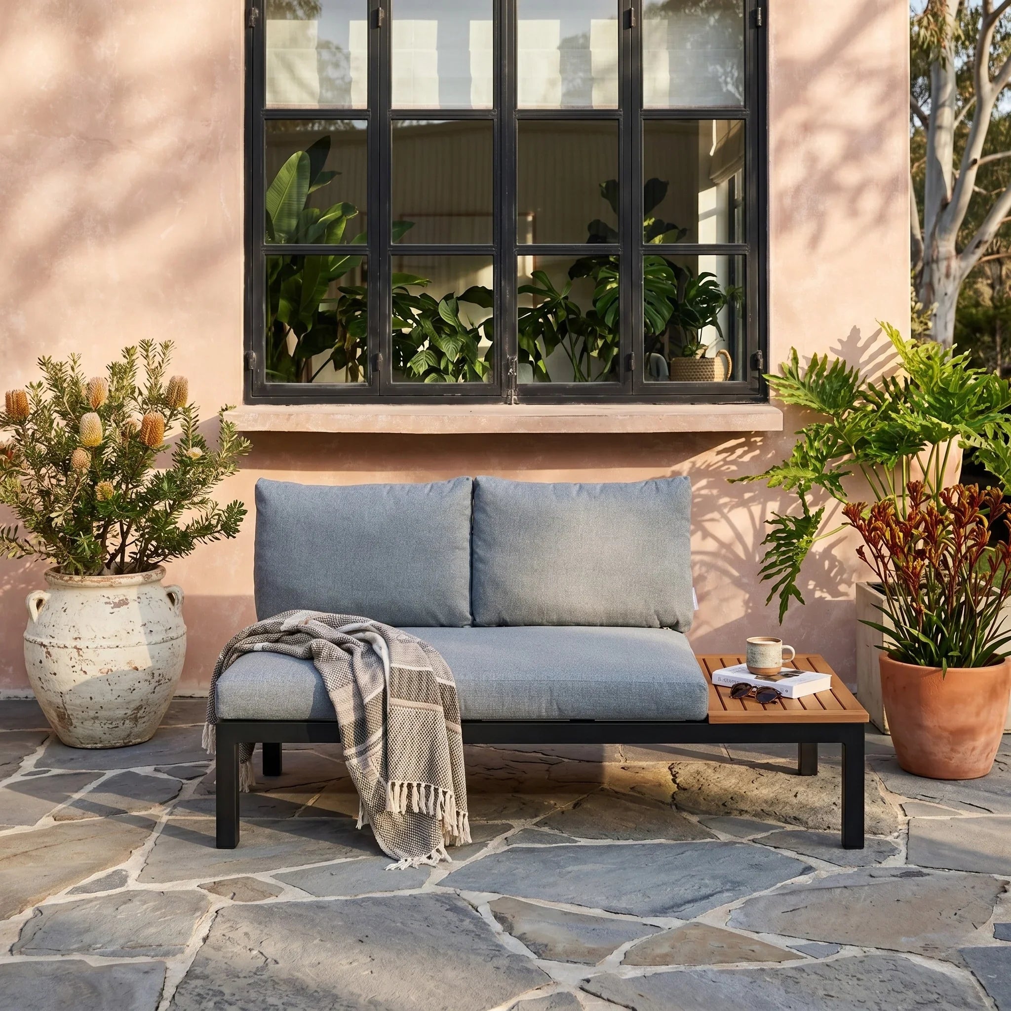 Outdoor patio setting with a blue cushioned sofa, potted plants, and a stone floor.