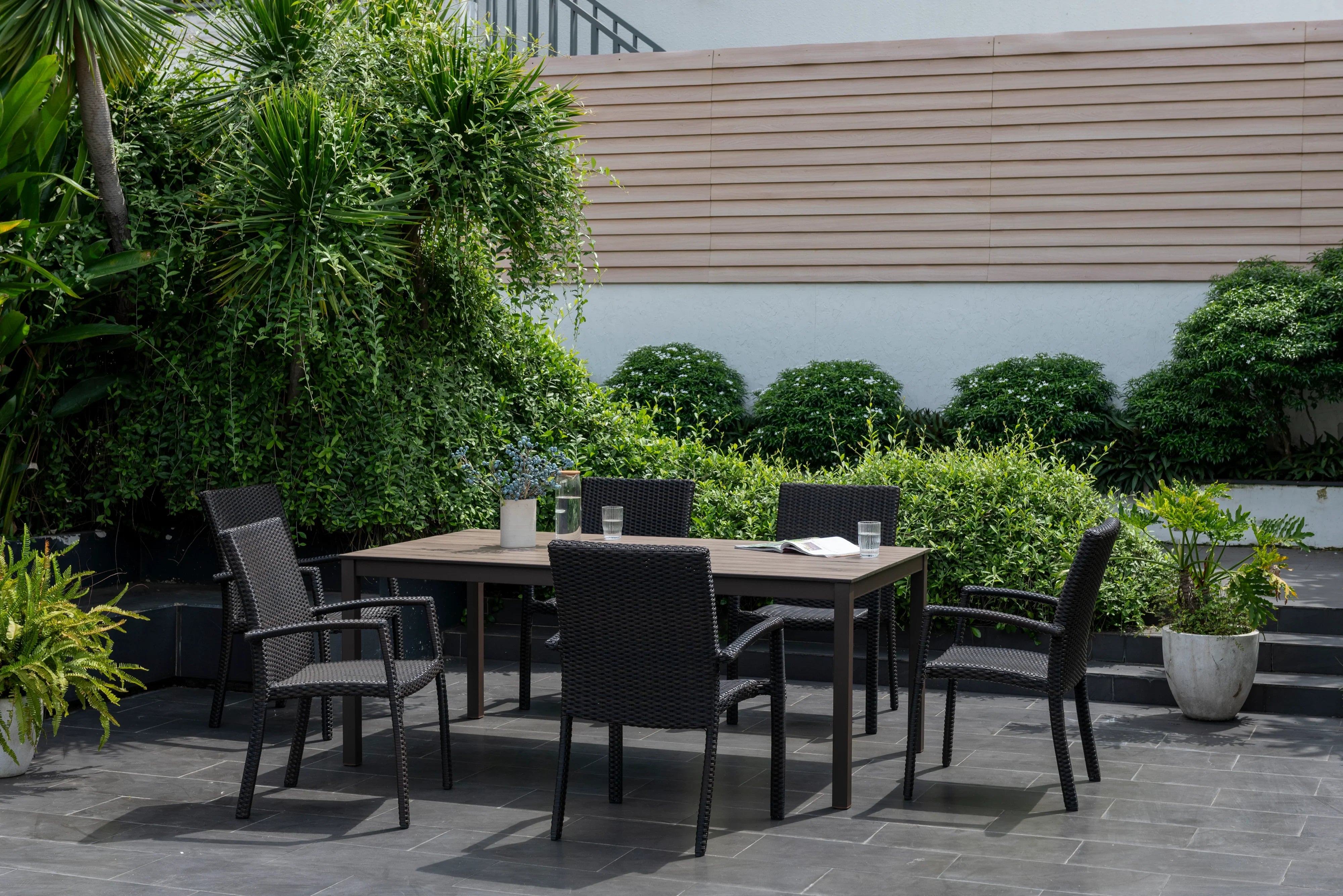 Outdoor dining set with six black wicker armchairs and rectangular table on patio, surrounded by greenery