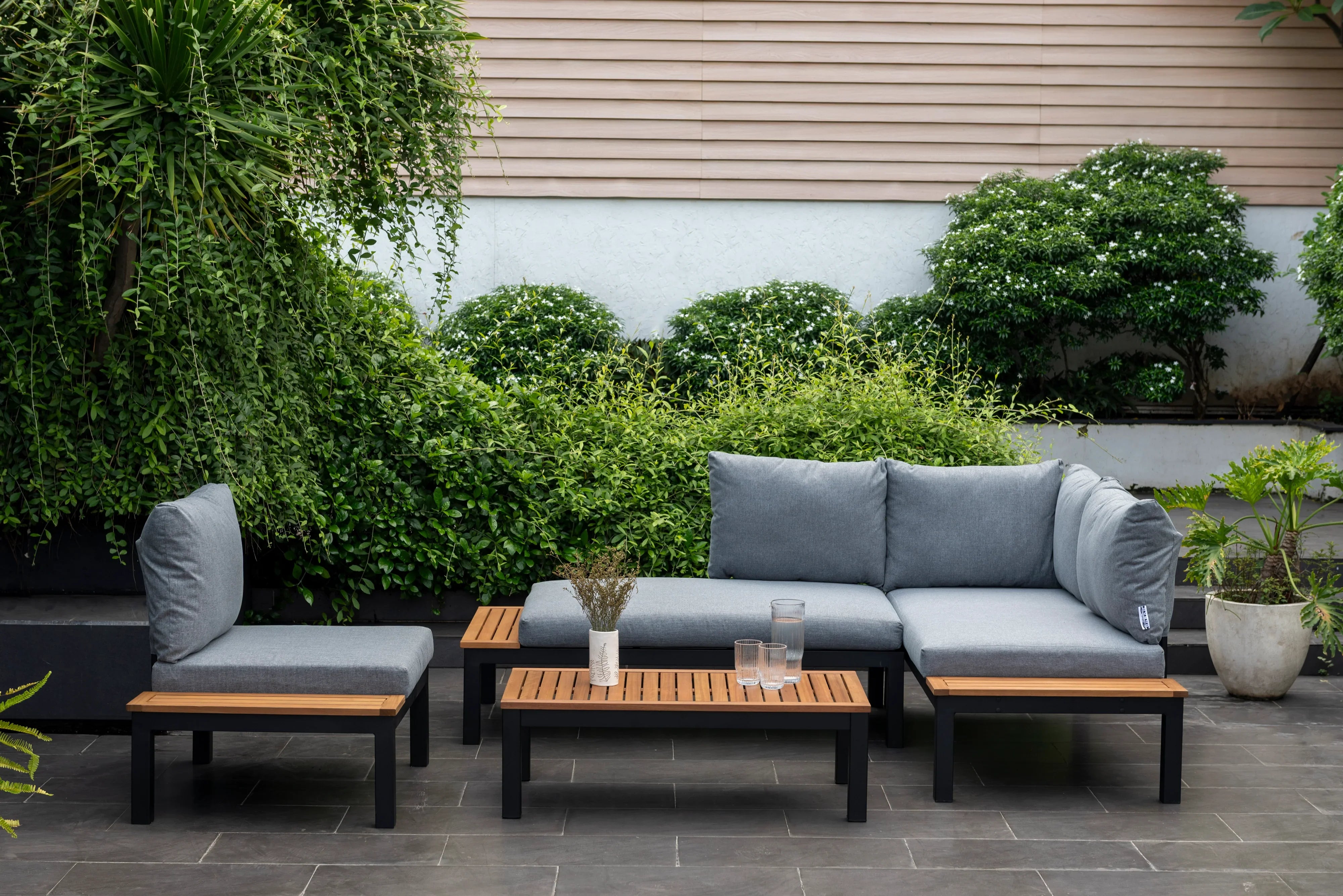 Modern outdoor sofa set with grey cushions and wooden accents on a patio, surrounded by green plants.