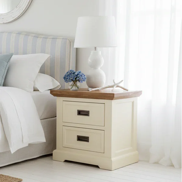 Two-drawer cream bedside table with wooden top, lamp, blue flowers, and coastal decor in bedroom.