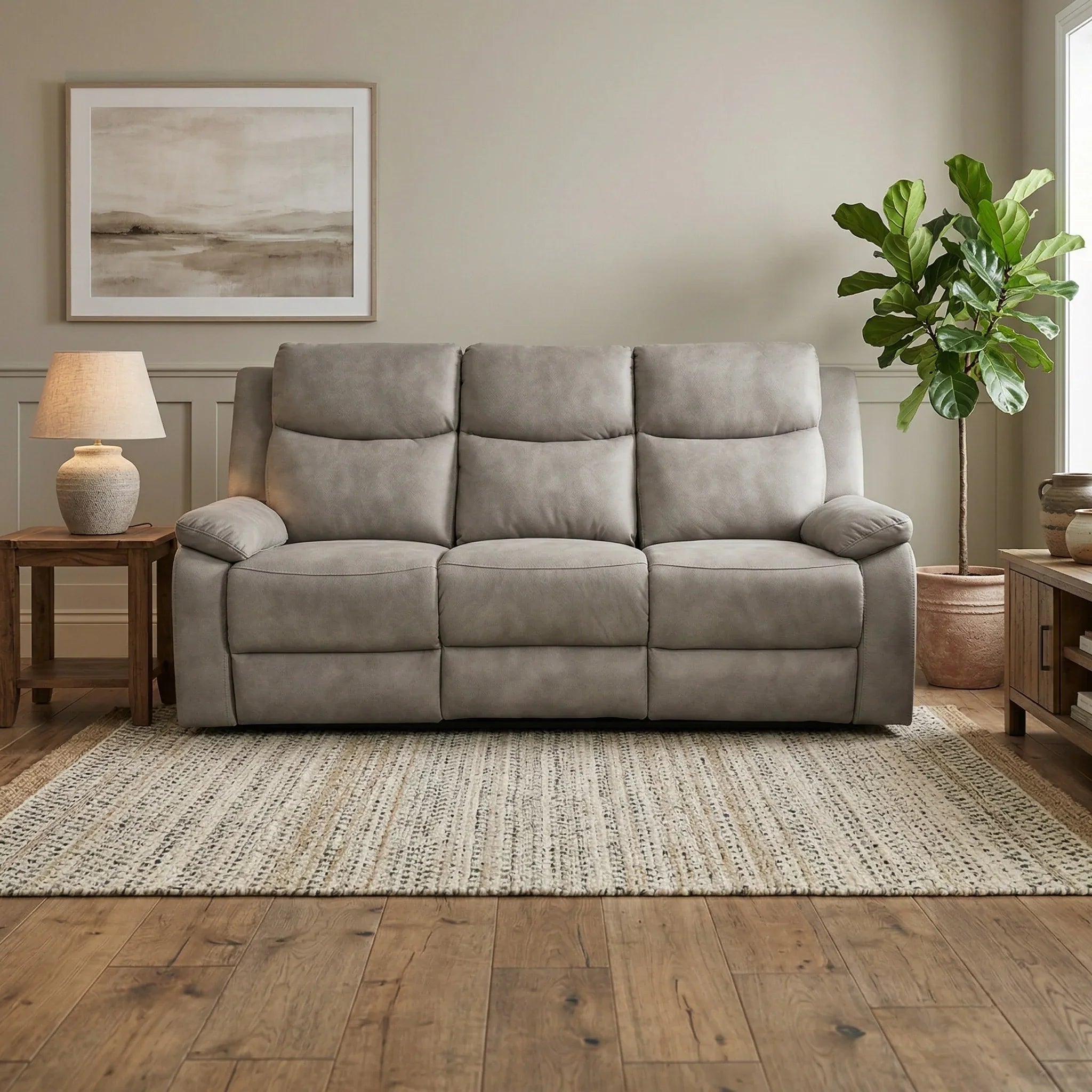 Mitcham 3 Seater Recliner Lounge in light grey with plush cushions and armrests, placed in a cozy living room setting with wooden furniture, a lamp, artwork, and a potted plant.