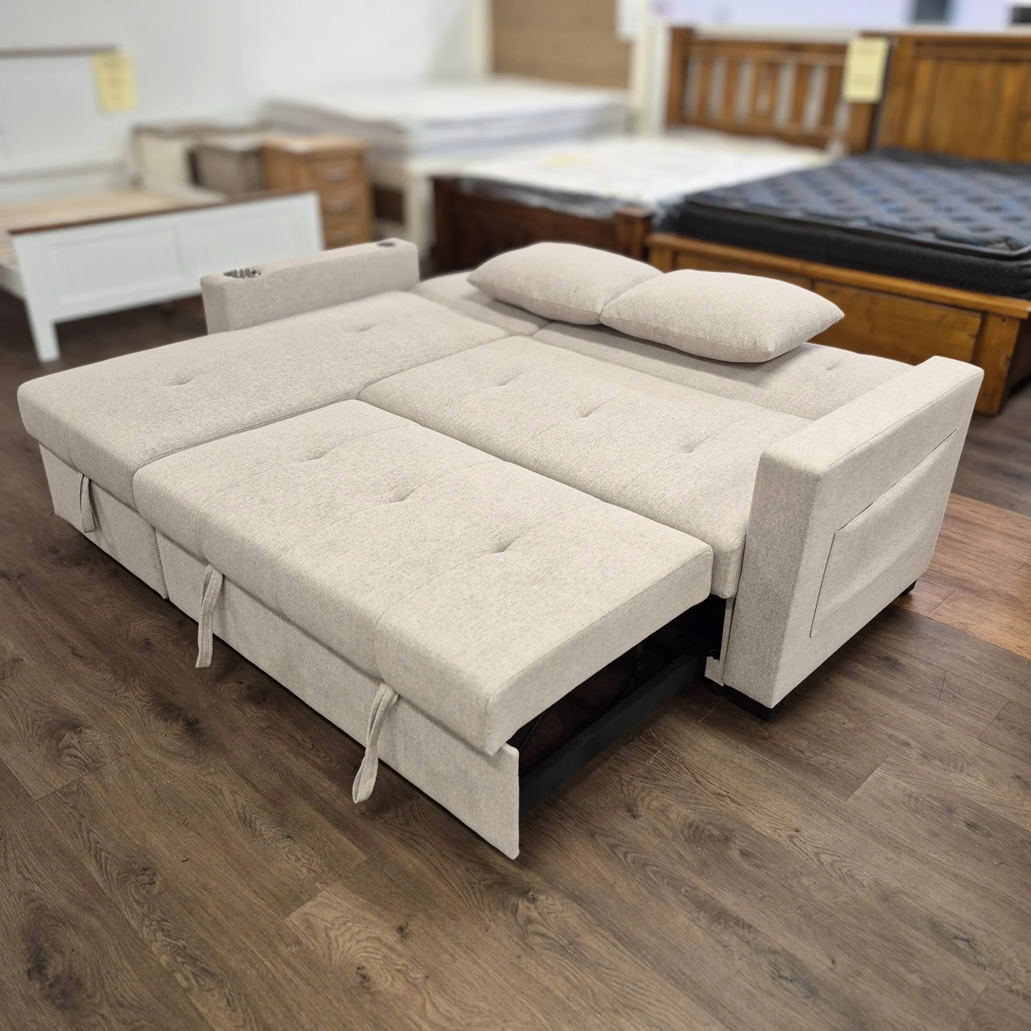 Beige sofa bed with tufted upholstery, extended flat with pillows, on wood floor in showroom