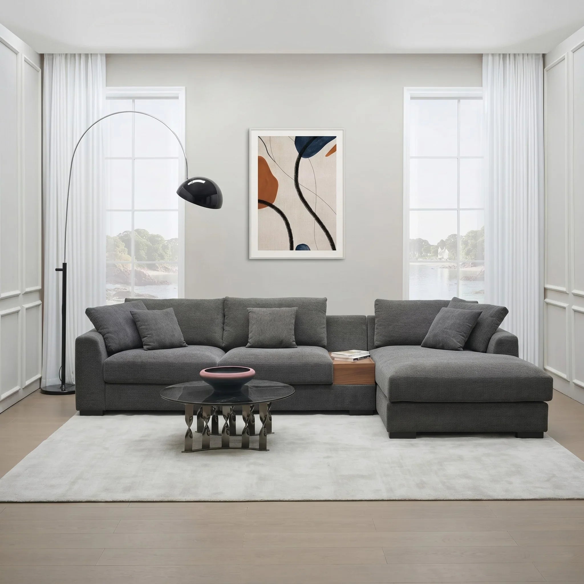 Odessey 3 Seater Chaise Sofa in dark gray fabric with multiple cushions, featuring a modern L-shaped design, placed in a bright living room with minimalist decor.