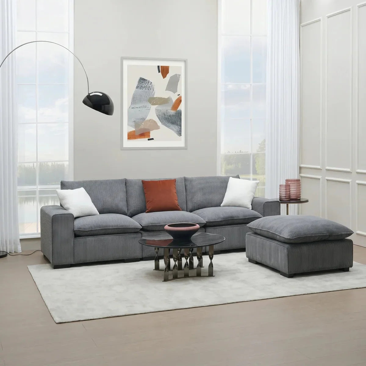 Orchid 3 Seater Sofa with Ottoman in a modern living room featuring soft gray upholstery, accented with white and orange cushions, paired with a round glass coffee table and floor lamp.