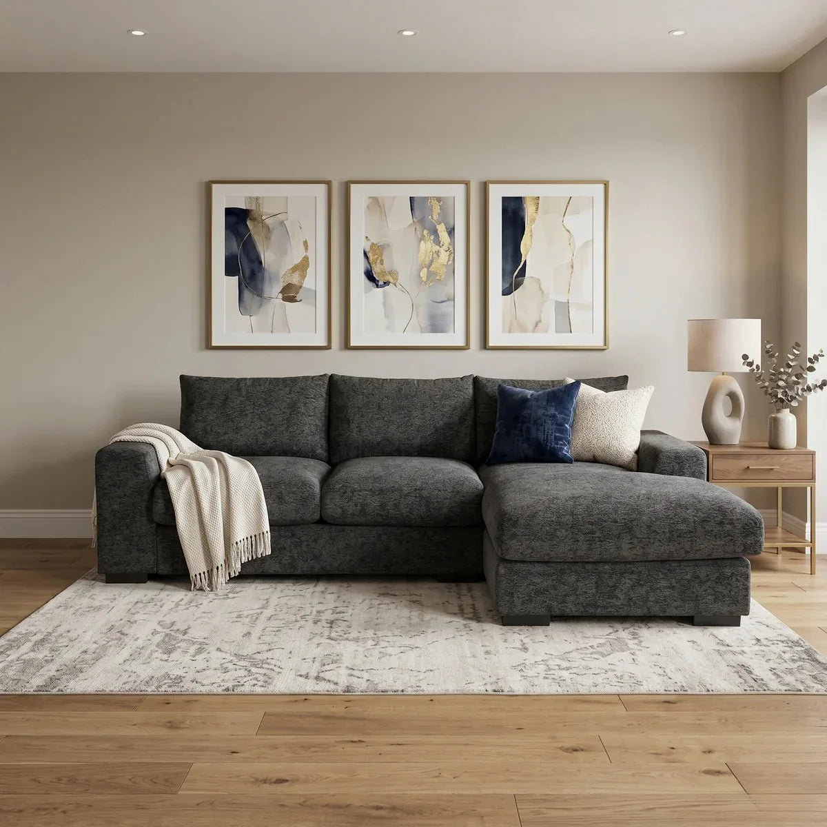 Oxford 3 Seater Couch with Chaise upholstered in dark grey fabric, styled with throw pillows and a blanket in a modern living room setting with abstract wall art and wooden flooring.