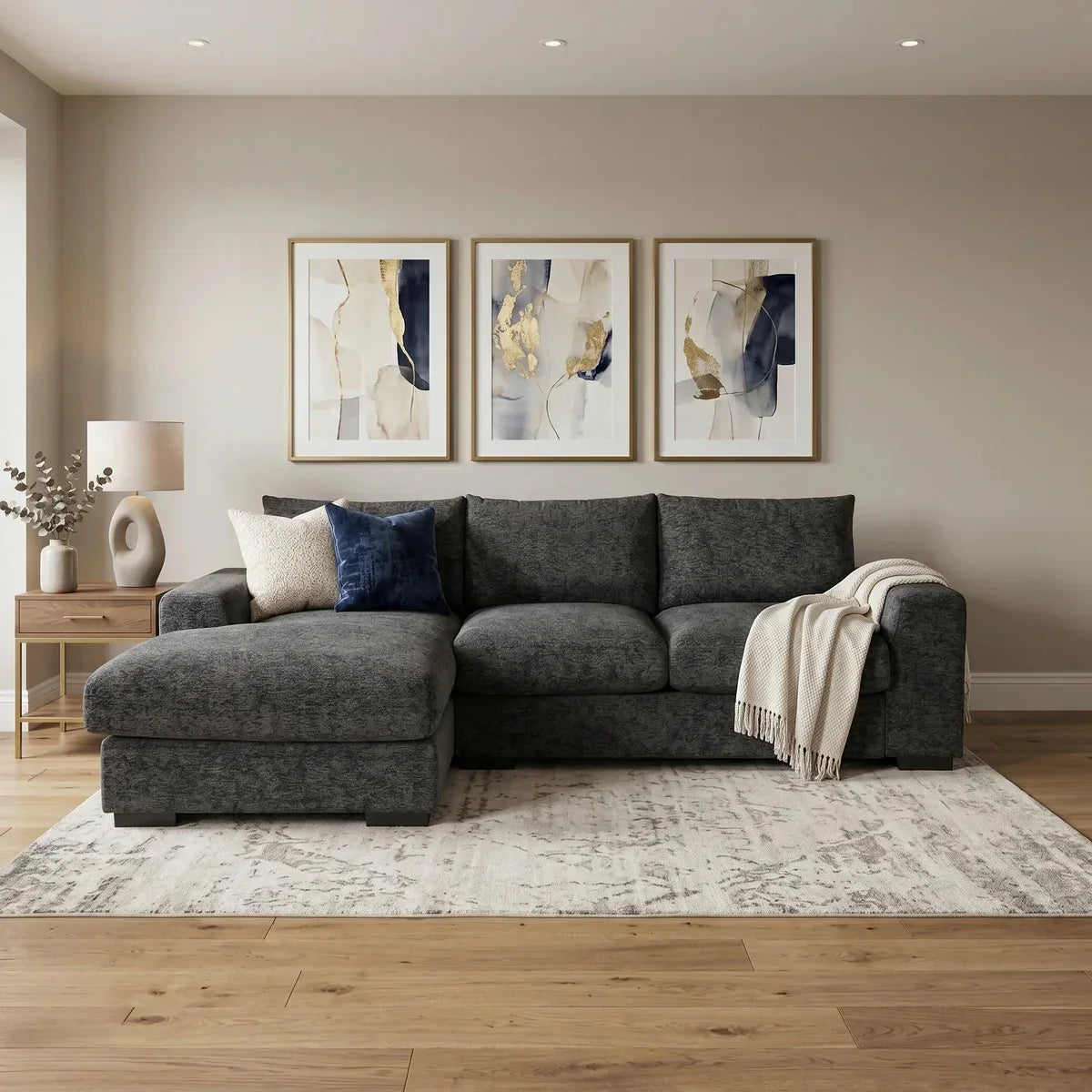Oxford 3 Seater Couch with Chaise in a modern living room setup featuring dark gray upholstery, decorative pillows, a throw blanket, wooden side table with lamp, and abstract wall art.