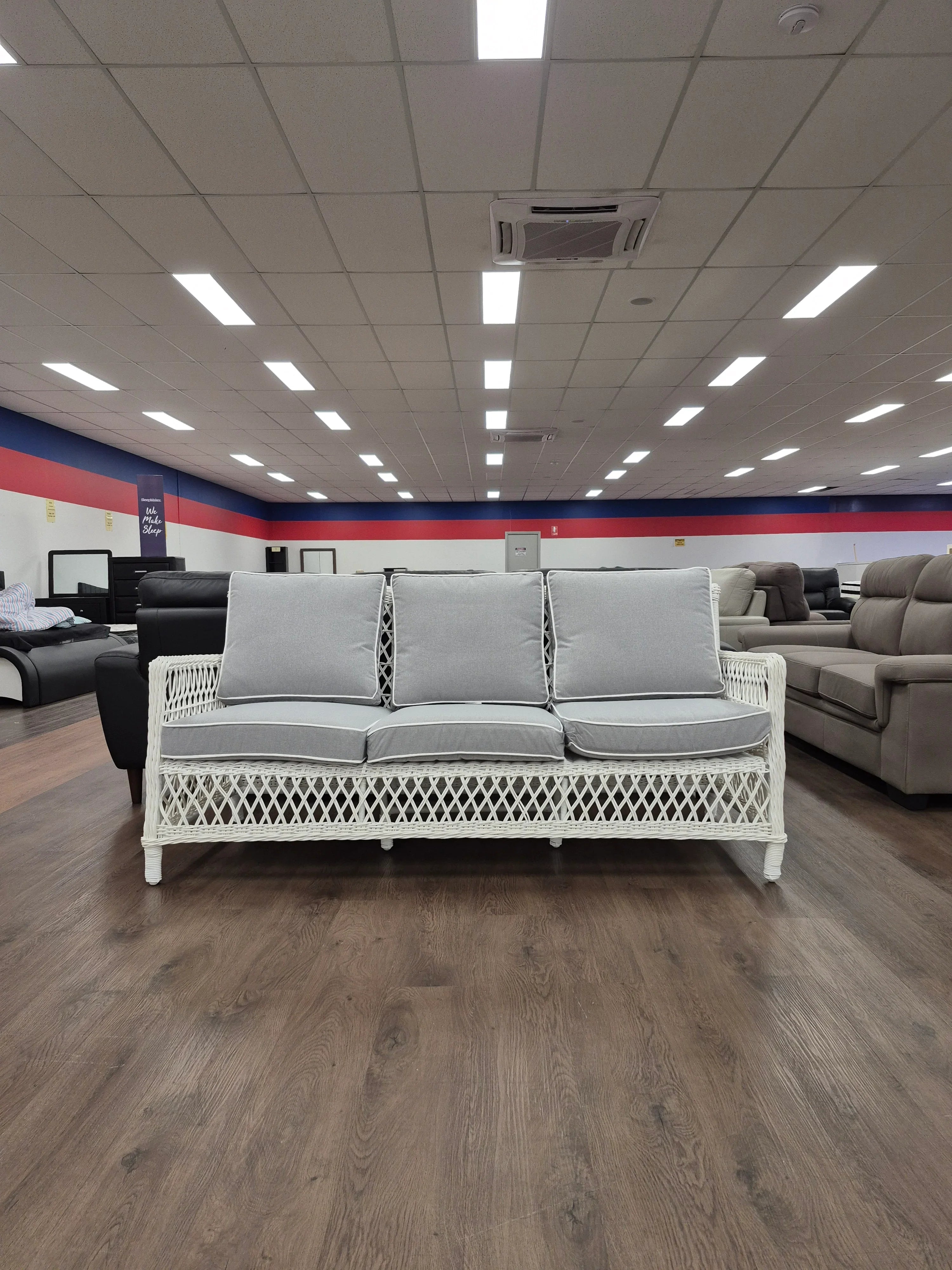 White wicker three-seat sofa with grey cushions in a furniture showroom on wood flooring