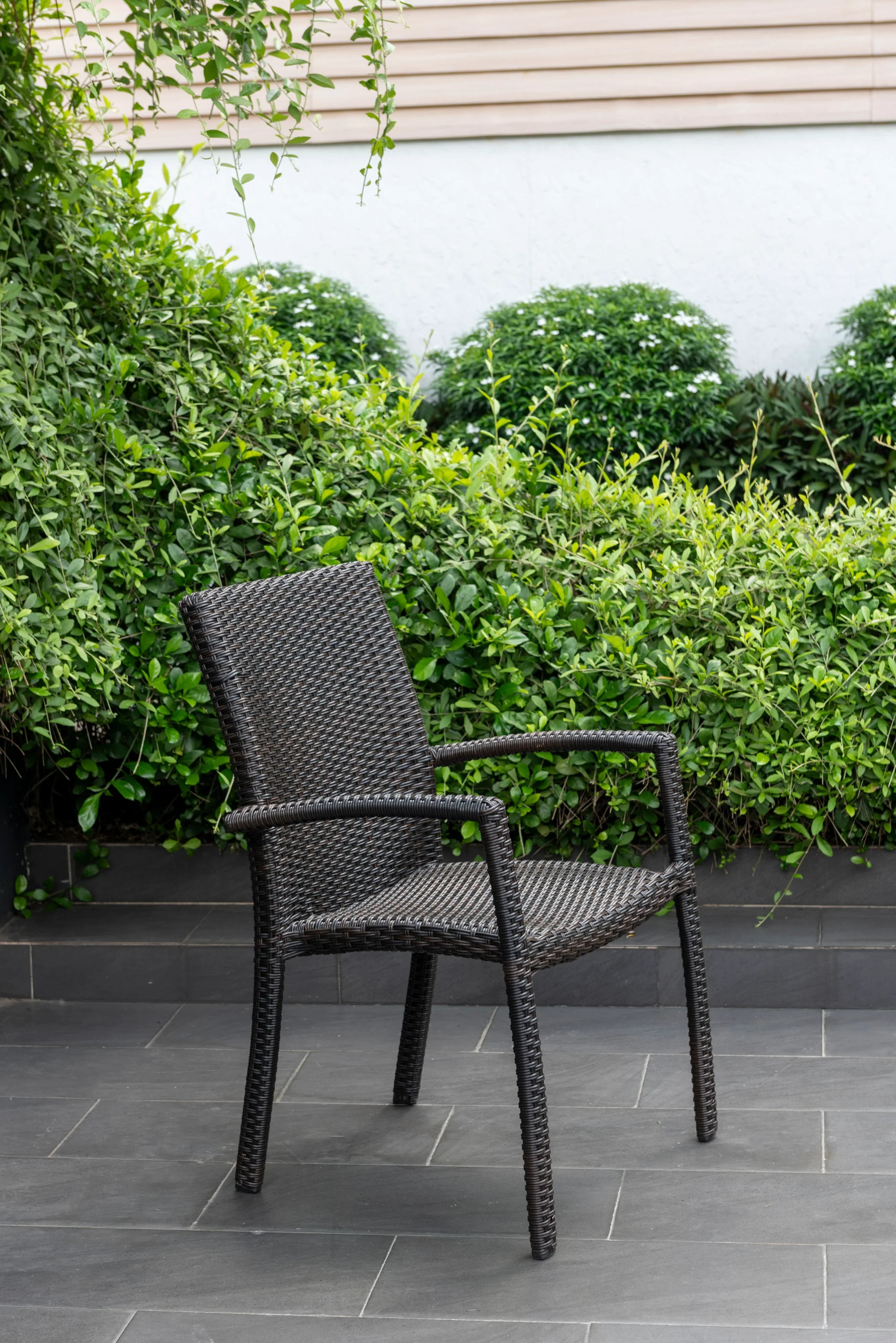 Outdoor rattan stacking armchair with armrests on tiled patio, surrounded by green bushes