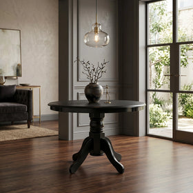 Daliya Round Dining Table in black with a turned pedestal column and carved four-leg base, shown in a modern room beside garden doors - The A2Z Furniture