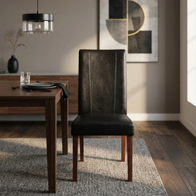 Dexter Chair in black leather-look upholstery with dark legs, shown against a concrete wall beside a timber dining table