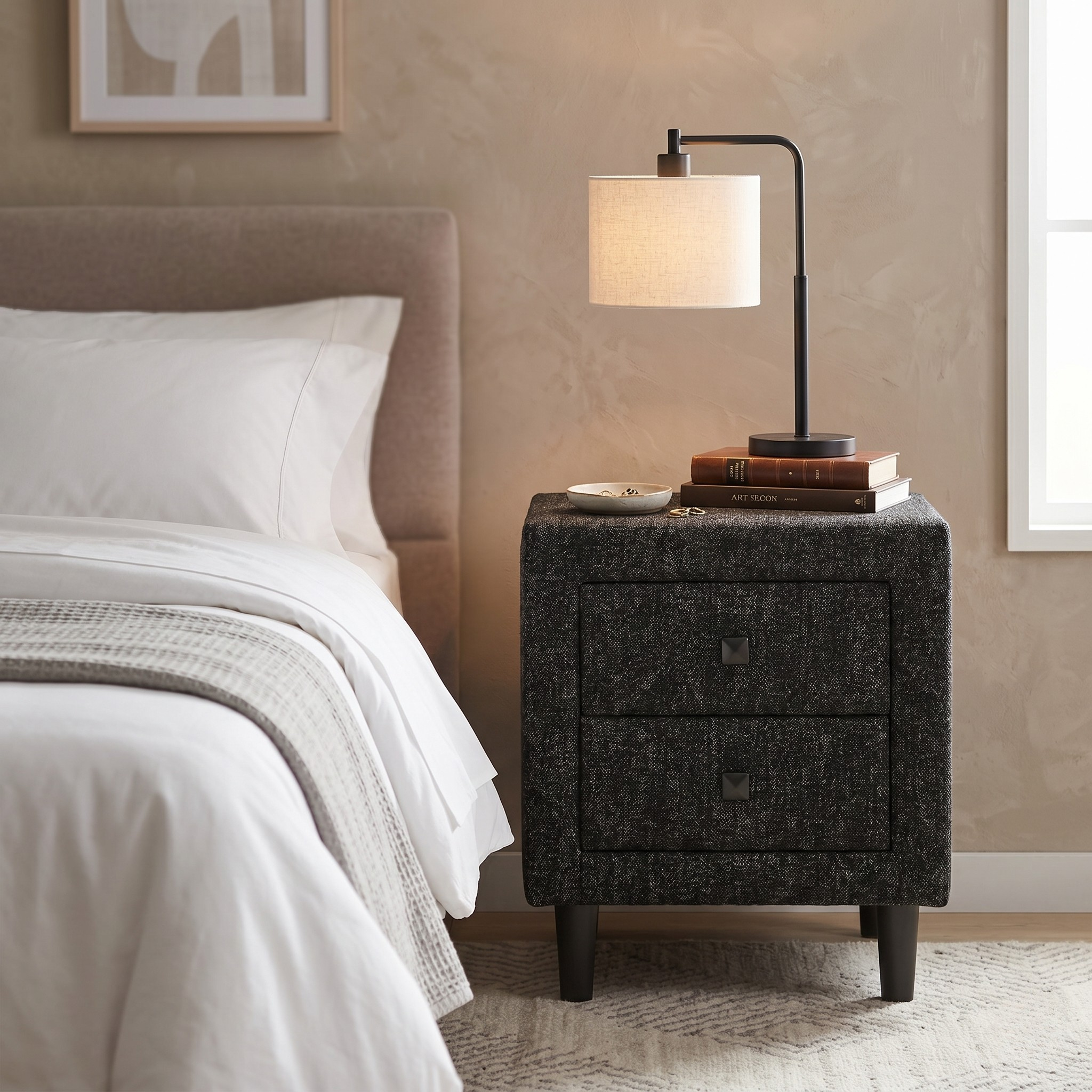Bedroom with a nightstand featuring a lamp and books, next to a bed with white bedding.