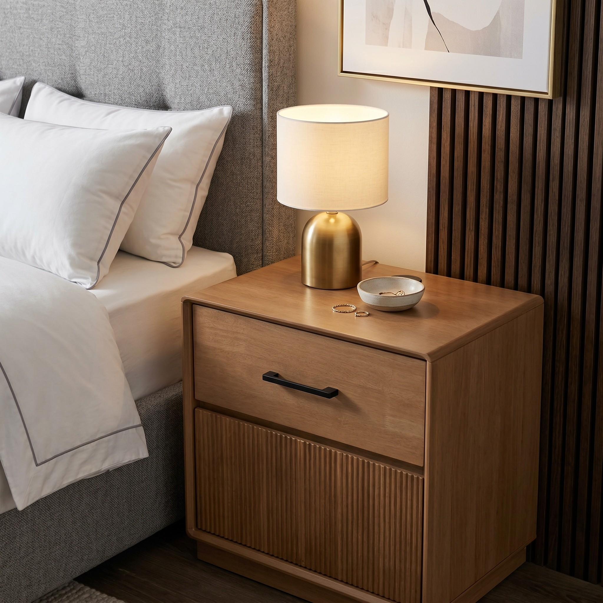 Wooden nightstand with a lamp and small bowl next to a bed in a bedroom setting.