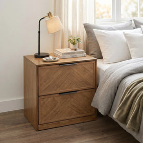 Wooden nightstand with lamp and books next to a bed with pillows and blanket.
