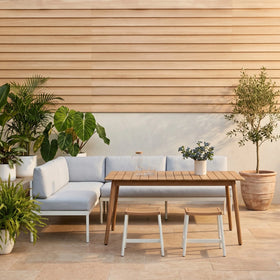 Bryde 4 Piece Outdoor Set featuring a modern L-shaped lounge with light cushions, a wooden coffee table, and two matching stools, styled in a bright outdoor patio with potted plants.