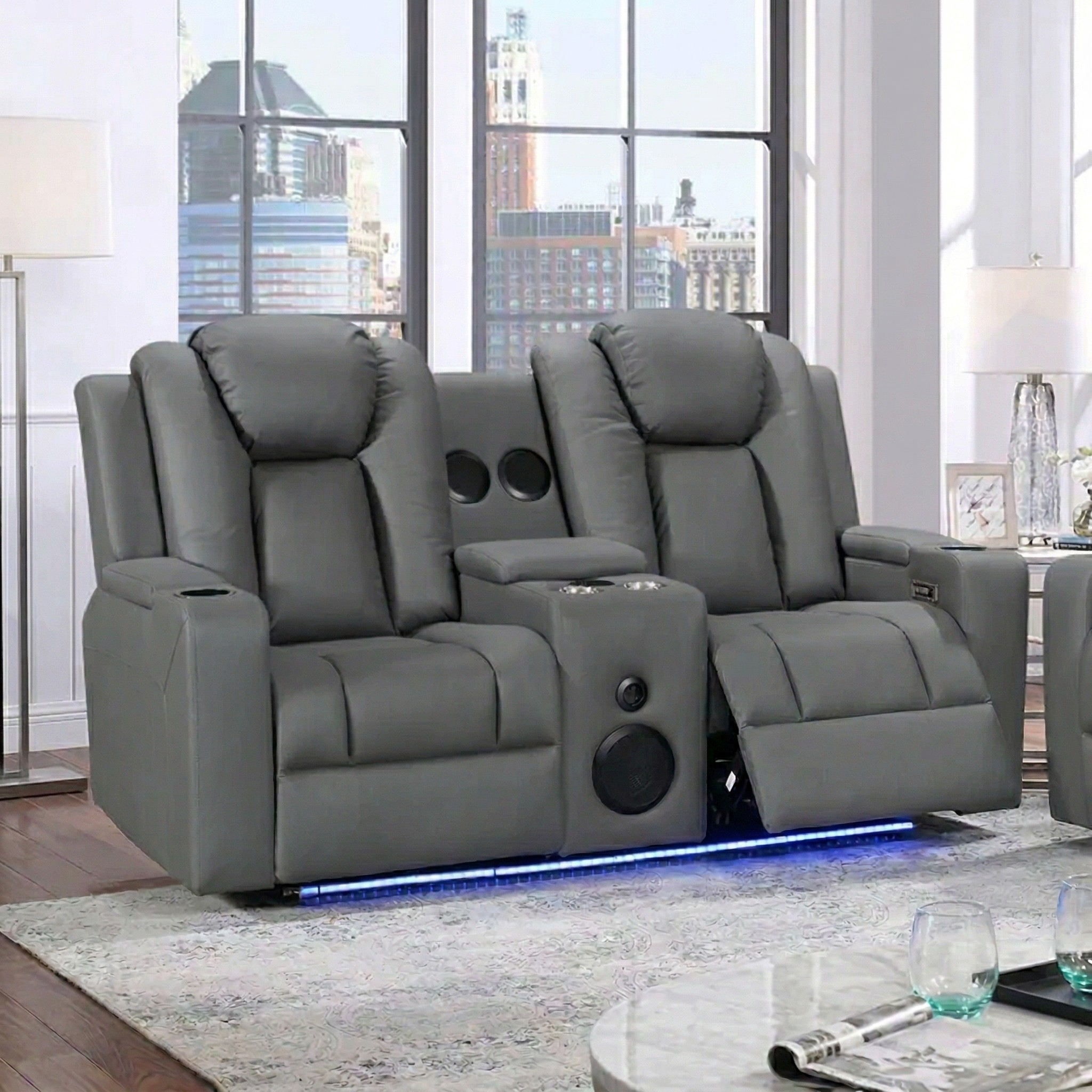 Breeze 2 Seater Dual Motor Electric Recliner Lounge in grey leather with built-in speakers and cup holders, set in a modern living room with cityscape windows.