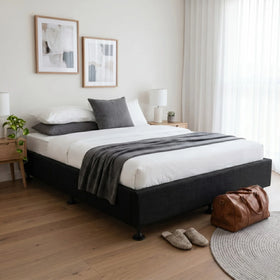 Modern bedroom showcasing the Ark Bed Base with a black fabric bed frame, neatly made white and gray bedding, paired with wooden side tables, lamps, and soft natural light.