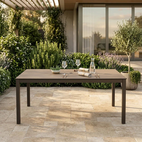 Wooden outdoor dining table with glasses and bottles on a patio with greenery and a building in the background