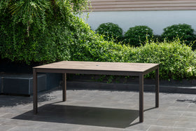 Rectangular outdoor dining table with slatted wood-look top on a tiled patio, garden background.