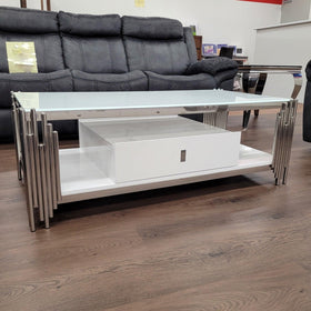 Modern glass-top coffee table with chrome legs and white storage drawer in showroom setting