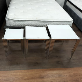 Modern glass top nest of tables set with metallic legs on wood floor near a bed