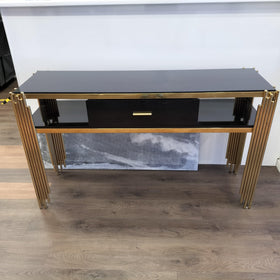 Black glass console table with gold metal legs and a central drawer, displayed indoors on wood flooring.
