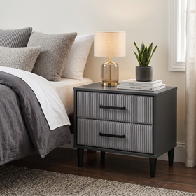 Frankston bedside table in dark grey with vertical fluted drawer fronts and black handles, topped with a lamp, books and a plant