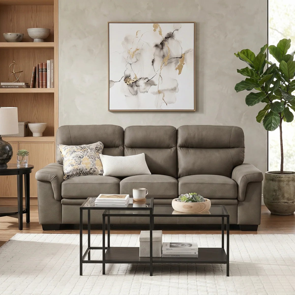 Erika 3 Seater Fabric Sofa in a neutral grey tone placed in a modern living room setting with a black metal coffee table, decorative cushions, indoor plant, and abstract wall art.