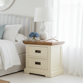 Two-drawer cream bedside table with wooden top, lamp, blue flowers, and coastal decor in bedroom.