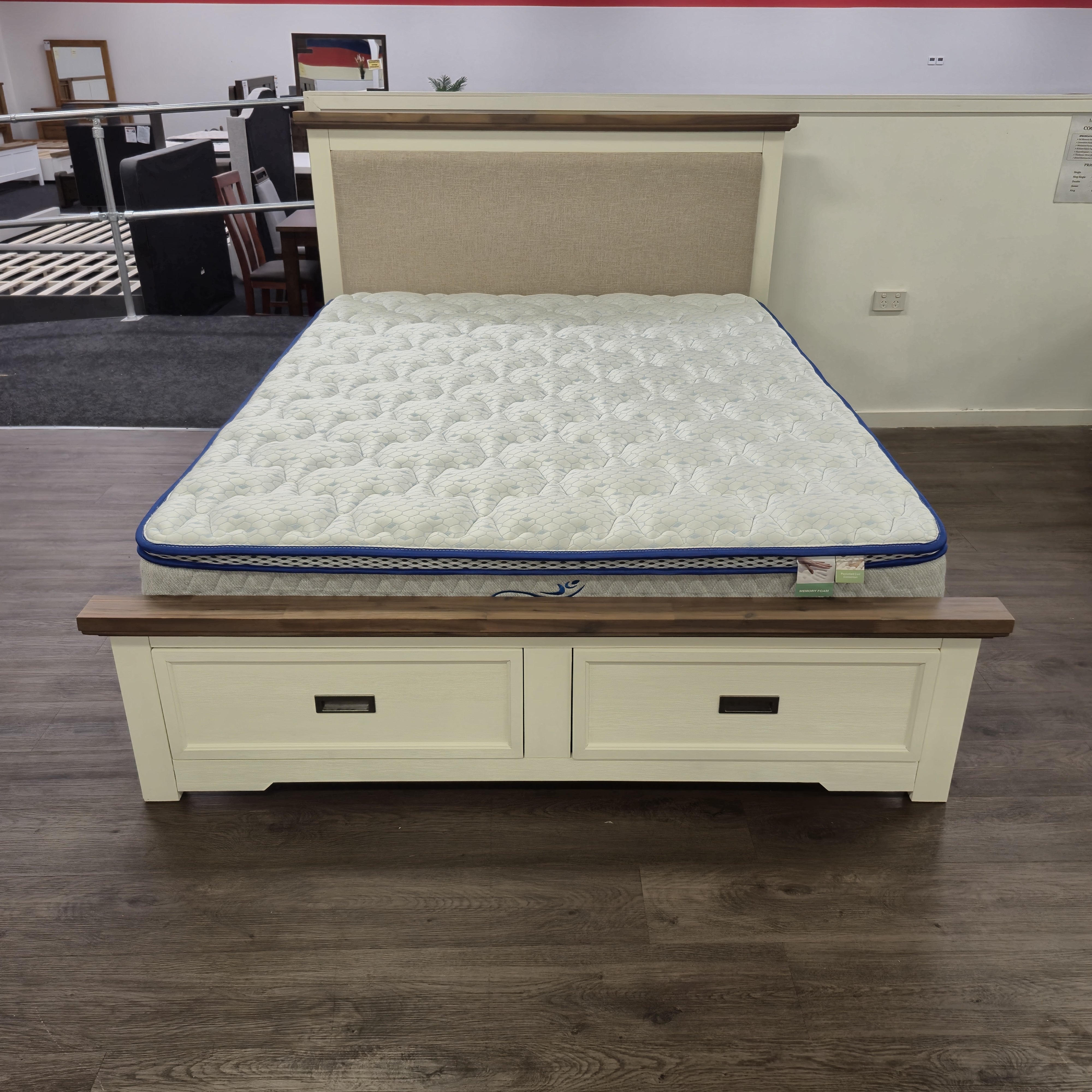 Jericho II Bed Frame with Storage, three-quarter angle view showing cream painted timber frame, walnut-toned wood top rails and oatmeal upholstered headboard panel, with a mattress on top, in The A2Z Furniture showroom