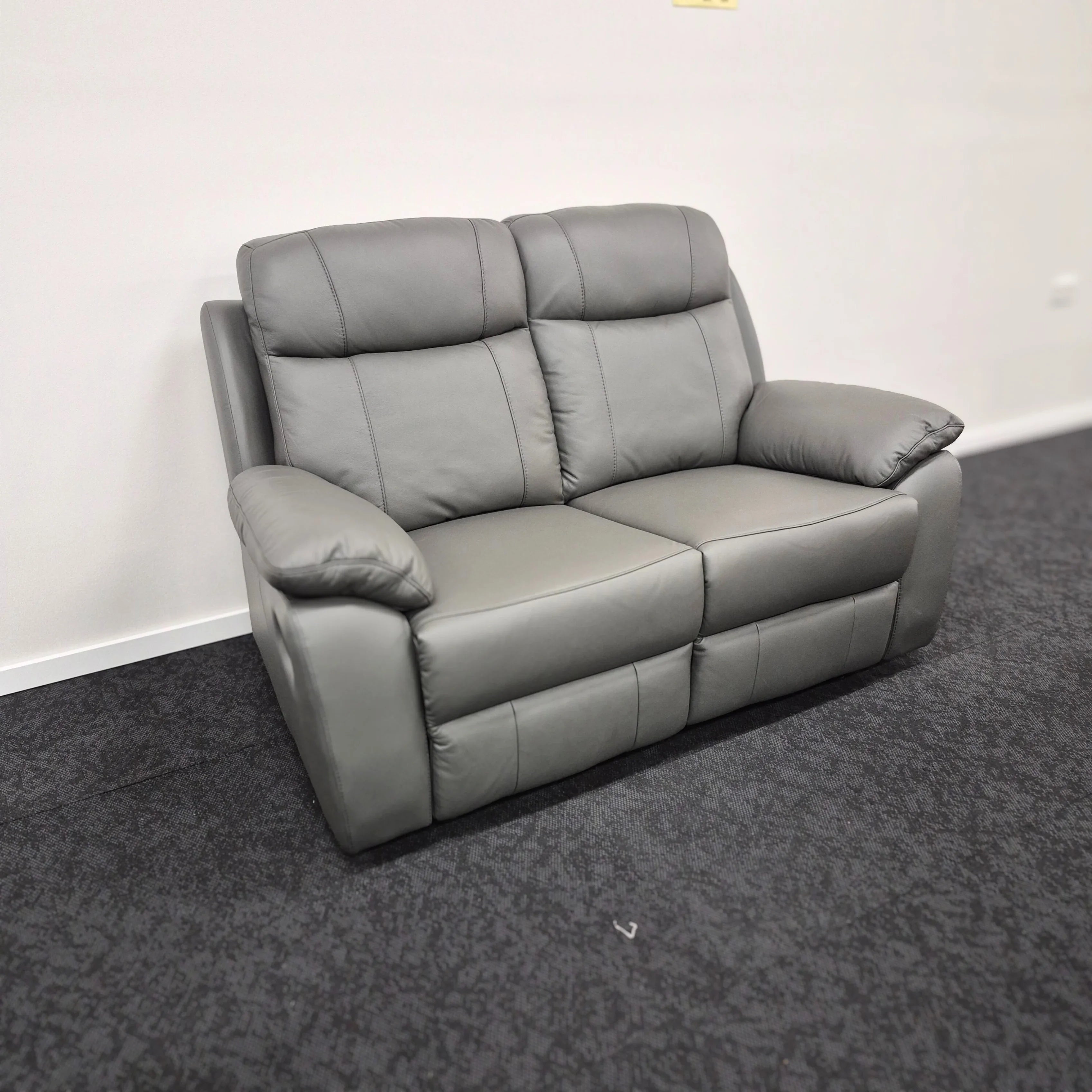 Grey leather 2-seater recliner sofa with padded armrests on dark carpeted floor