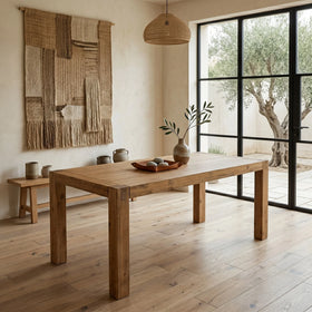 Wooden dining table in a room with large windows and a textured wall hanging.