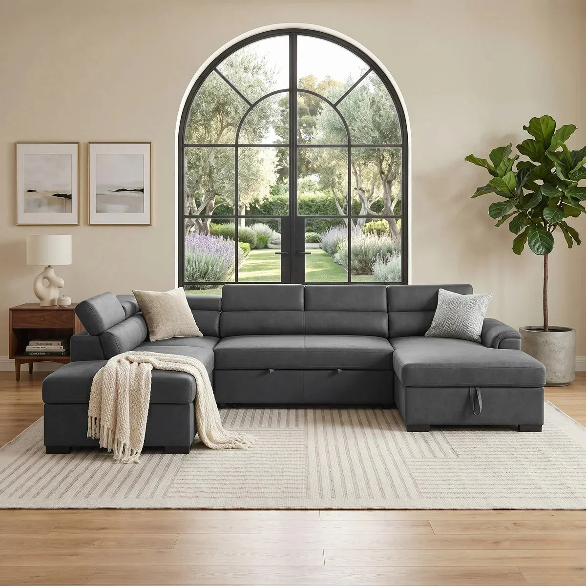 Spacious modern living room featuring the Omaha Pull-Out Sofa Bed in dark gray fabric with plush cushions, decorative pillows, a throw, and a large arched window overlooking a garden.