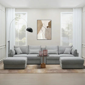 Modern living room featuring a light gray Oracle 4 Seater Sofa with Ottoman, complemented by neutral tones, large windows, minimalist decor, and a sleek floor lamp.