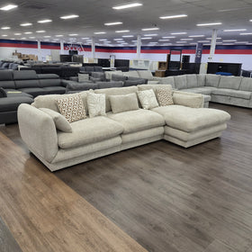 Otway L-Shaped Fabric Lounge in Beige viewed from a raised angle in the showroom, showing the chaise depth and scatter cushions.