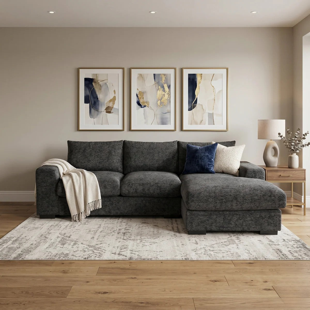 Oxford 3 Seater Couch with Chaise upholstered in dark grey fabric, styled with throw pillows and a blanket in a modern living room setting with abstract wall art and wooden flooring.
