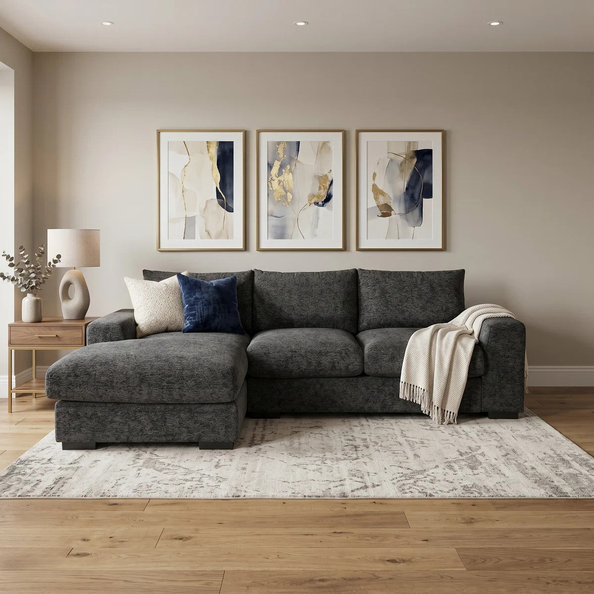 Oxford 3 Seater Couch with Chaise in a modern living room setup featuring dark gray upholstery, decorative pillows, a throw blanket, wooden side table with lamp, and abstract wall art.