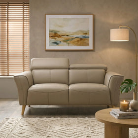 Penelope 2 Seater Leather Lounge in beige with soft cushions and wooden legs, styled in a cozy living room with modern decor including a floor lamp, wall art, and textured rug.