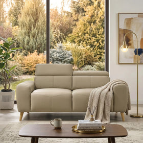 Percy 2 Seater Leather Lounge in beige with modern wooden legs, styled in a cozy living room setting featuring a throw blanket, coffee table with books and cup, a floor lamp, and large windows overlooking autumn trees.