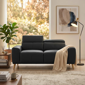 Percy 2 Seater Leather Lounge in black with wooden legs, set in a cozy living room with a beige throw, floor lamp, abstract wall art, and a side table with books and decor.