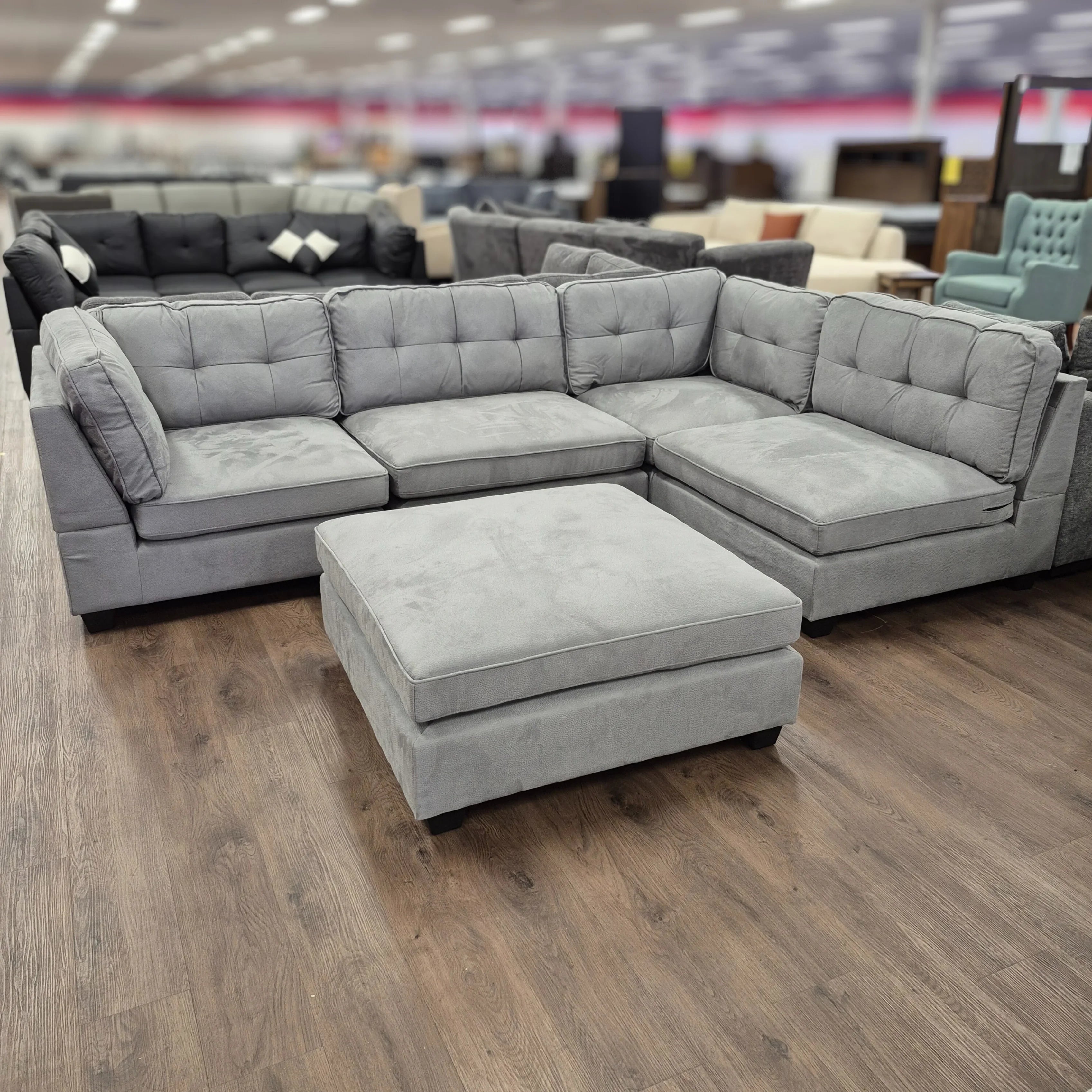 Grey corner sectional lounge with ottoman, tufted cushions in showroom on wood floor